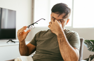 Tired man takes off his glasses and rubs his eyes while reading a book at home.