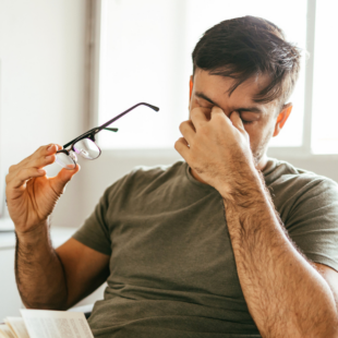 Tired man takes off his glasses and rubs his eyes while reading a book at home.