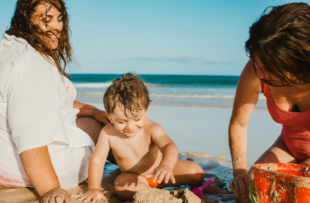 Adults smiling and watching cute boy building castle from wet sand while resting on beach near sea together