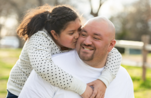 Daughter hugging her dad from behind and kissing his cheek.