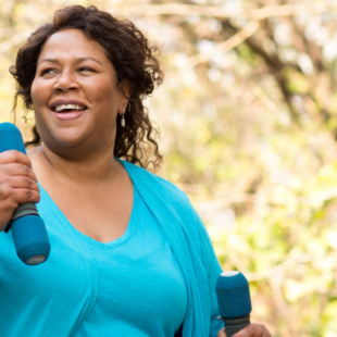 A woman walking in a park with two light dumbbells.