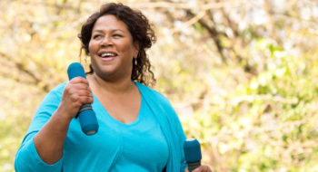 A woman walking in a park with two light dumbbells.