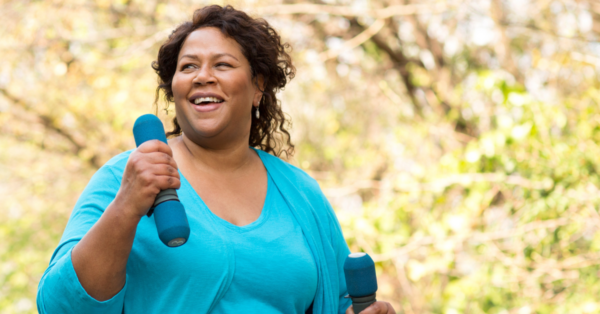 A woman walking in a park with two light dumbbells.