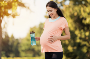 Pregnant woman in a pink t-shirt is standing in the park with a sports bottle in hands.