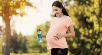 Pregnant woman in a pink t-shirt is standing in the park with a sports bottle in hands.