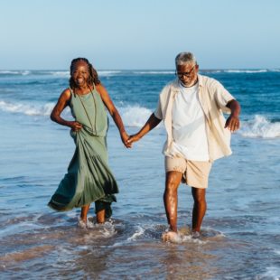 A cheerful older couple holds hands while walking and laughing together along the sandy beach, embodying joy and togetherness.