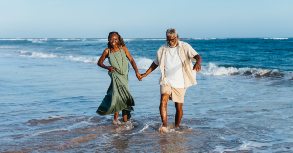 A cheerful older couple holds hands while walking and laughing together along the sandy beach, embodying joy and togetherness.