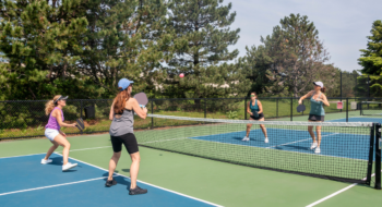 A comptetivie doubles game of pickleball at the net on a blue and green court in summer.