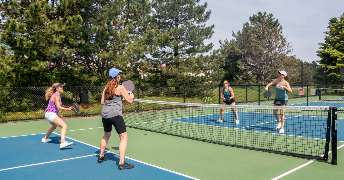 A comptetivie doubles game of pickleball at the net on a blue and green court in summer.