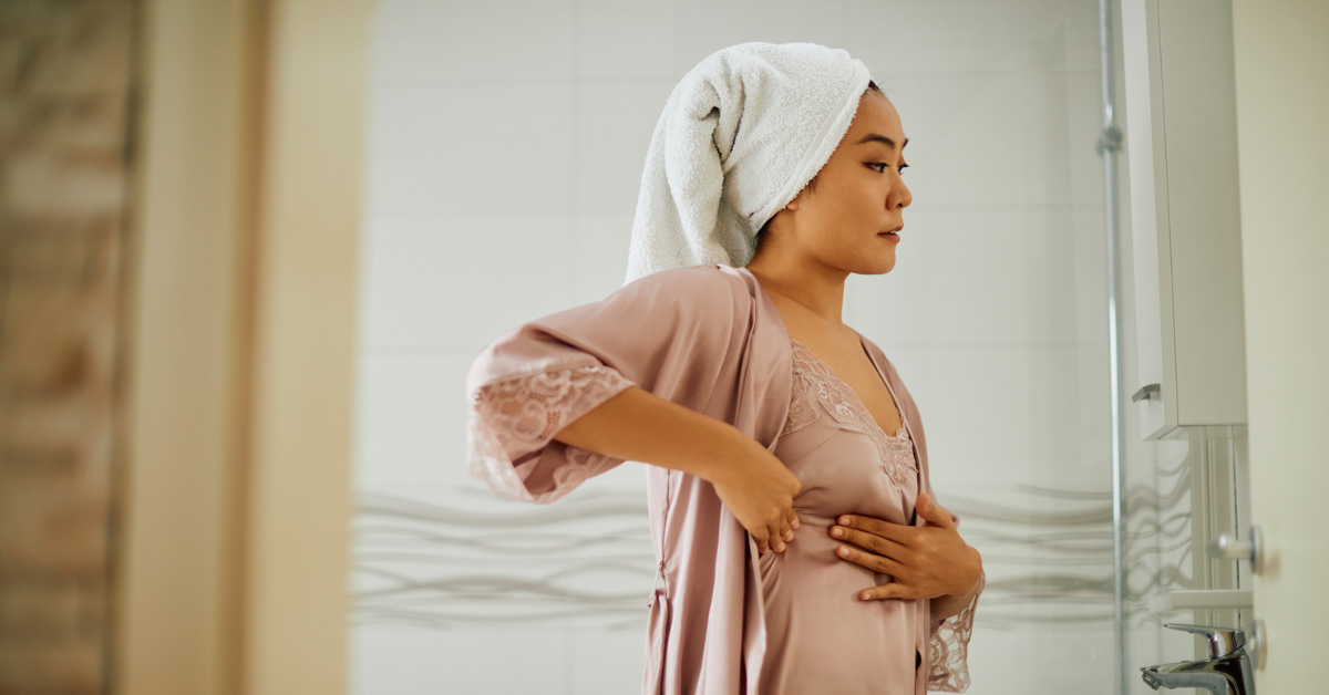 Young woman standing in front of bathroom mirror while doing breast self-examination.