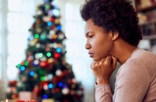 A woman sits with her head resting on her hands while looking concerned or worried. There is a Christmas tree lit up in the background and she appears to be at home.