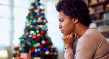 A woman sits with her head resting on her hands while looking concerned or worried. There is a Christmas tree lit up in the background and she appears to be at home.