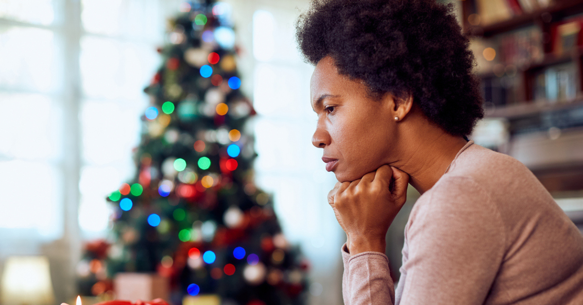 A woman sits with her head resting on her hands while looking concerned or worried. There is a Christmas tree lit up in the background and she appears to be at home.