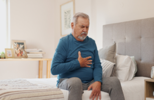 Senior man grabbing his chest area while sitting on the edge of the bed, suffering with acid reflux.