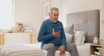 Senior man grabbing his chest area while sitting on the edge of the bed, suffering with acid reflux.