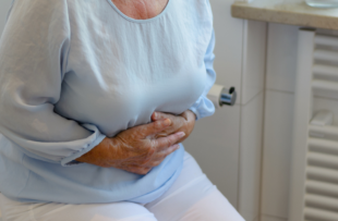 Mid section of senior woman wearing white trousers and light blue blouse sitting on toilet holding stomach.