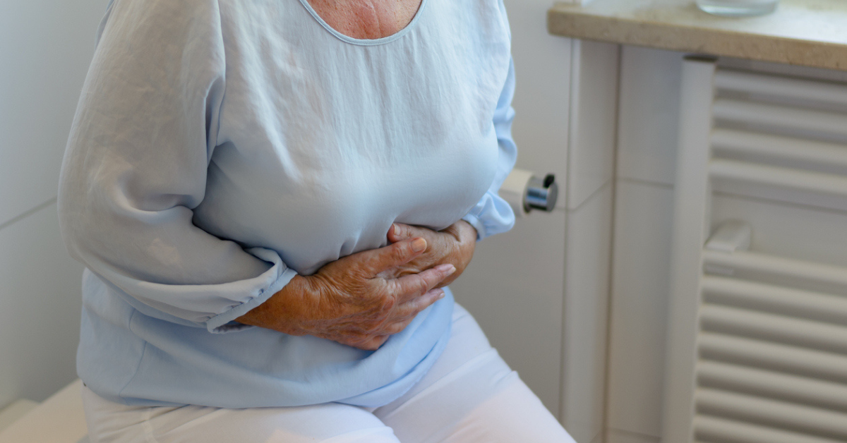 Mid section of senior woman wearing white trousers and light blue blouse sitting on toilet holding stomach.