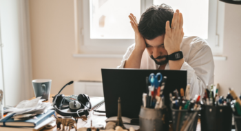 A man sitting at his desk with his computer rubbing his head in frustration.