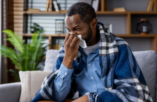 A man sits on his couch wrapped in a blanket and blowing his nose.