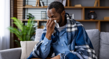 A man sits on his couch wrapped in a blanket and blowing his nose.