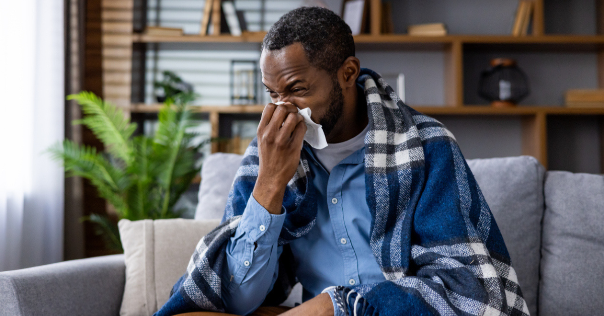 A man sits on his couch wrapped in a blanket and blowing his nose.