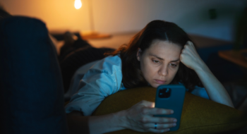 Young serious sad woman lying on sofa and looking at smartphone screen at night or evening